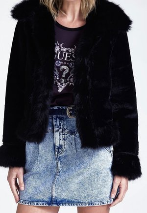 Femme portant une veste en fausse fourrure noire sur un t-shirt sombre à logo, associée à une mini-jupe en jean délavé bleu clair et une ceinture.