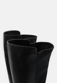 Botas de cuero negro hasta la rodilla con una textura suave, bordes redondeados y amplias aberturas en la parte superior. Diseño minimalista sin herrajes visibles.