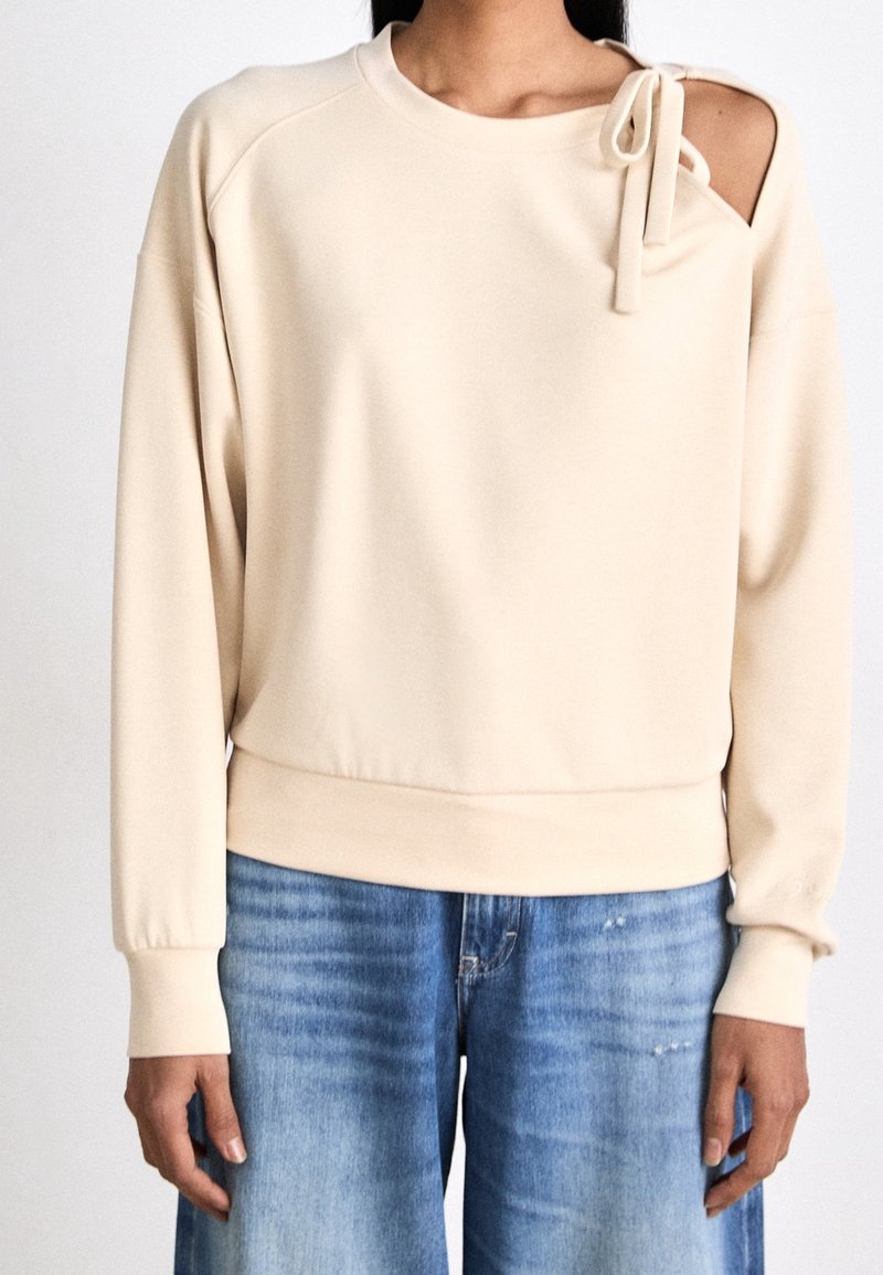 Sweat-shirt beige à manches longues avec une découpe et un nœud noué sur l'épaule gauche, porté avec un jean en denim bleu.