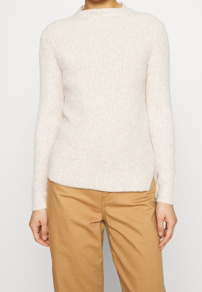 Personne portant un pull en maille côtelée blanc et un pantalon marron clair, visible des épaules jusqu'aux hanches sur un fond uni.
