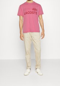 Rosa bomull t-shirt med "CLUB LACOSTE" tryckt i mörkare rosa, kombinerad med beige chinos och vita sneakers. Enkel design med rundad hals.