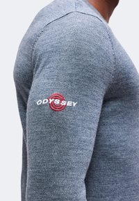 Gros plan d'une manche de chemise grise à manches longues avec un logo "ODYSSEY" brodé en rouge et blanc sur le haut du bras.