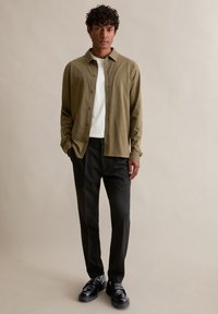 Olijfgroen overhemd met knopen over een wit t-shirt, gecombineerd met zwarte broek en zwarte schoenen, staand tegen een neutrale achtergrond.