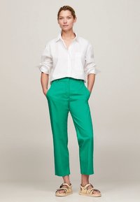 Hvit skjorte med knappelukking, stukket inn i livlige grønne bukser med en rettskåret design. Modellen har på seg beige sandaler med svarte detaljer.