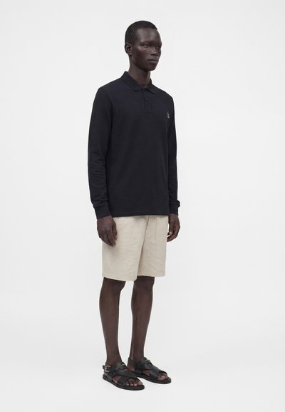 Homme portant un polo noir à manches longues, un short beige et des sandales noires, debout devant un fond blanc uni.