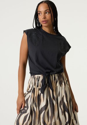Zwart katoenen top met een geknoopt voorpand, versierd met gelaagde bloemrijke borduursels. Gecombineerd met een geplooide rok in beige en donkerbruine strepen.