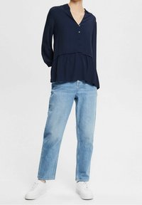 Blouse à manches longues bleu marine avec un col en V, des boutons à l'avant et un ourlet à volants, associée à un jean droit bleu clair et des baskets blanches.