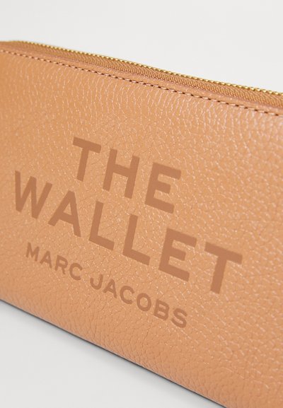Portafoglio in pelle marrone chiaro con finitura testurizzata, chiusura con zip dorata e testo stampato in evidenza: "IL PORTAFOGLIO MARC JACOBS."