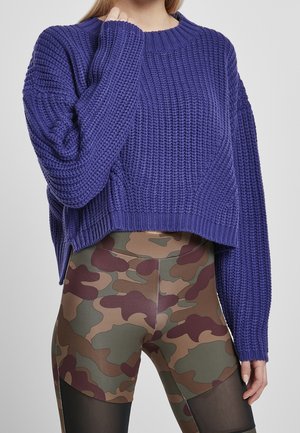 Personne portant un pull en tricot violet et des leggings à motif camouflage avec des panneaux en maille noire, debout devant un fond uni.