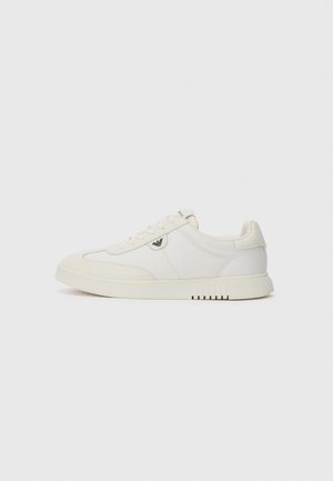Baskets blanches montantes avec lacets, semelle lisse et petit logo métallique sur le côté, présentées sur un fond uni.