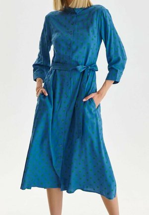 Blauwe midi-jurk met groene stippen, knoopsluiting aan de voorkant, driekwart mouwen, tailleband met strik en handen in de zakken.