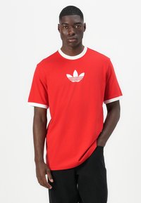 Κόκκινο βαμβακερό T-shirt με λευκά περιθώρια στους καρπούς και τον λαιμό. Διαθέτει το λευκό λογότυπο της Adidas στο στήθος. Κλασική γραμμή, κοντά μανίκια.