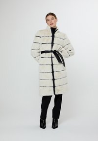 Bílý kabát z umělé kožešiny s černými pruhy, s vysokým límcem, zipovým zapínáním a černým páskem jako akcentem. Nositý přes černé kalhoty a boty.