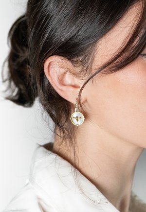 Guldtonade örhängen med en rund vit emaljpendant med ett bivedesign. Texturerade accenter och en hängare i form av en ring ger extra detaljer.