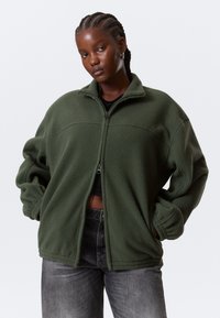 Femeie cu păr împletit purtând o jachetă de fleece verde măsliniu peste un top negru și blugi închiși la culoare decolorați, stând pe un fundal alb.