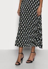 Svart plisserad midikjol med ett geometriskt diamantmönster i blått och vitt, matchad med svarta höga sandaler med remmar.