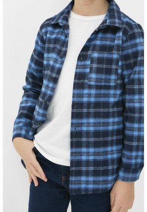 Dziecko ubrane w niebiesko-czarną kratowaną koszulę z długim rękawem na białej koszulce i ciemnoniebieskie dżinsy, stojące z jedną ręką w kieszeni.