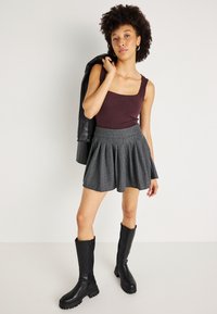 Μπορντό μπλούζα χωρίς μανίκια, γκρι ψηλόμεση φούστα με πτυχές και ρίγες, μαύρες μπότες μέχρι το γόνατο με χοντρές σόλες, μοντέλο holding μια ζακέτα.