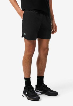 Homme portant un short de sport noir, des chaussettes noires et des baskets Lacoste noires, debout, les mains le long du corps, sur fond blanc.