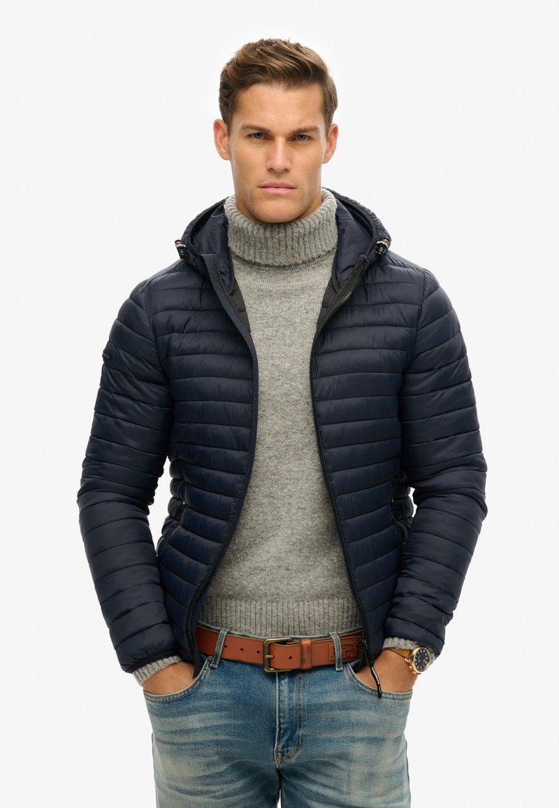 Marineblå, kviltet pufferjakke med hætte, båret over en lys grå turtleneck sweater. Modellen har hænderne i lommerne, iført denim jeans og et brunt bælte.