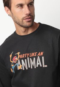 Camisa negra de algodón de manga larga con un diseño gráfico de un león de dibujos animados y el texto "FIESTA COMO UN ANIMAL" en colores contrastantes.