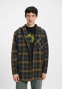 Sort og orange ternet skjorte med hætte, med knaplukning foran og en brystlomme, båret over en sort grafisk t-shirt.