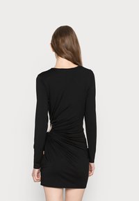 Vestido preto de mangas longas com um design justo, apresentando um detalhe de tecido torcido na parte de trás e um recorte lateral para um toque adicional.