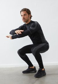 Svart träningsklädsel som inkluderar en åtsittande långärmad topp med texturerade ränder och matchande leggings. Modellen utför en knäböj. Svarta skor.