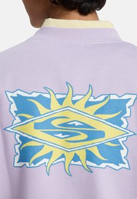 Μπλούζα με κουκούλα λιλά με κίτρινο αντίθετο γιακά. Διαθέτει ένα μεγάλο γραφικό στην πλάτη: έναν σχεδιασμό ήλιου με μπλε και κίτρινες αποχρώσεις.