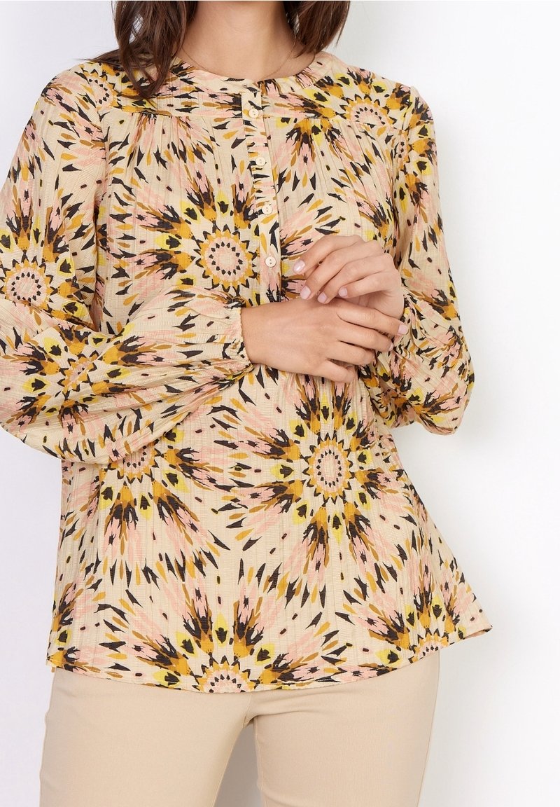 Blusa a maniche lunghe color crema con un vivace motivo floreale nei toni del giallo, rosa e nero. Presenta un colletto rotondo e dettagli con bottoni. Tessuto testurizzato.