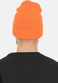 Orange strikket hue med en foldet kant, der har en blød tekstur og en spidse top. Båret af en person foran en neutral baggrund.