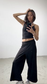 Fekete, rövidített tank top fodros szegéllyel és magas derekú, bő szárú fekete nadrággal. A modell fehér sportcipőt visel, és tükör előtt pózol.