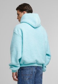 Light blue fleece hoodie with a textured, plush finish, featuring a large hood and ribbed hem. Pairs with blue denim pants.