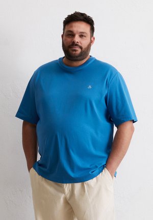 Blaues Baumwoll-T-Shirt mit rundem Ausschnitt und kurzen Ärmeln, das ein kleines, gesticktes Logo auf der Brust zeigt. Kombiniert mit beigen Hosen.