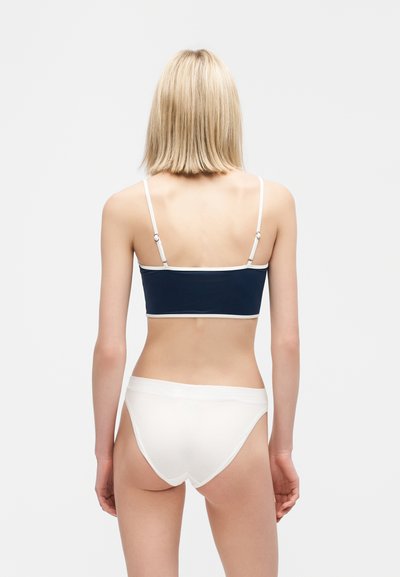 Γυναίκα με ξανθά μαλλιά που φοράει ένα μπλε σκούρο τοπ χωρίς τιράντες με λευκό πλαίσιο και λευκά κάτω μέρη μπικίνι, στέκεται μπροστά σε ομοιόμορφο φόντο.