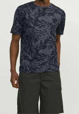 Uomo che indossa una maglietta blu navy a maniche corte con motivi scuri a foglia e pantaloncini scuri fino al ginocchio, in piedi davanti a uno sfondo uniforme.