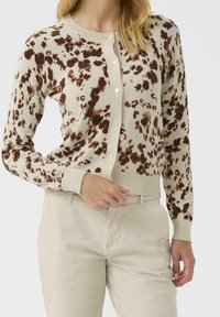 Cardigan beige avec des taches léopard brunes, présentant un col rond, des fermetures à boutons, ainsi que des poignets et un ourlet côtelés, associé à un pantalon de couleur claire.