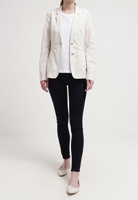 Blazer beige chiaro con due bottoni, abbinato a una blusa bianca con texture e jeans neri aderenti. Modella che indossa ballerine color crema.