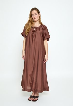 Femme aux longs cheveux portant une robe ample marron jusqu'aux chevilles et des sandales noires, debout devant un fond blanc uni.