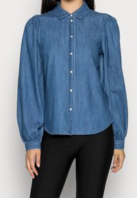 Mujer vestida con una blusa azul de mezclilla con botones de manga larga y hombros abullonados, junto con pantalones negros ajustados.