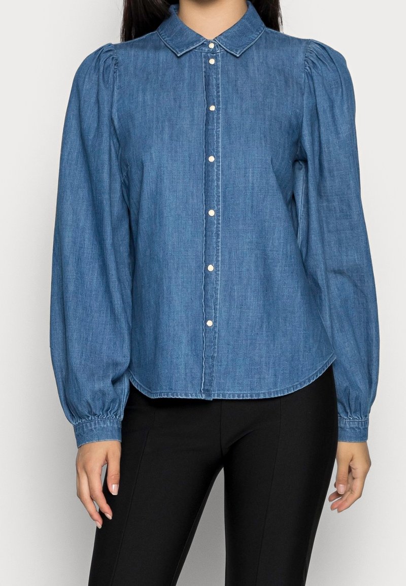 Mujer vestida con una blusa azul de mezclilla con botones de manga larga y hombros abullonados, junto con pantalones negros ajustados.