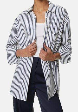 Persoon draagt een loszittend blauw-wit verticaal gestreept overhemd over een wit hemdje en marineblauwe broek, en houdt de randen van het overhemd vast.