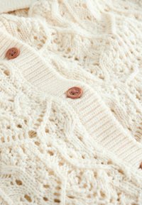 Tissu tricoté couleur crème avec des motifs complexes de torsades et de dentelle, doté d'une bande de boutons côtelée avec des boutons marron.