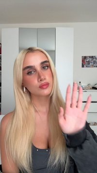 Lange, glatte blonde Haare umrahmen ein Gesicht mit heller Haut und definierten Augenbrauen. Sie trägt ein schwarzes Oberteil, eine Hand ist in einer Geste erhoben.