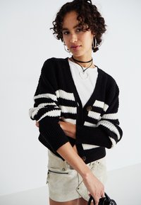 Black and white striped knitted cardigan with long sleeves, button closure, and textured fabric. Paired with a beige mini skirt.