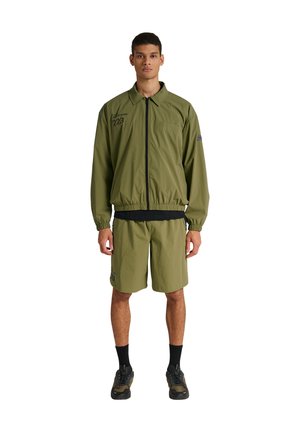 Mand står med fronten mod kameraet iført olivengrøn lynlåstrøje og matchende shorts, sorte sokker og mørke sneakers på hvid baggrund.