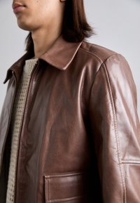 Mees, kes kannab pruuni nahkjakki kreemi kudumisega sviidri peal, näidates jope krae, lukku ja esi-tasku detaile.