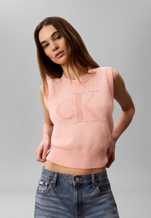 Jonge vrouw met lang bruin haar draagt een mouwloze roze gebreide top met "CK" logo en blauwe spijkerbroek, poserend tegen een grijze achtergrond.