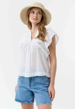 Woman wearing beige wide-brimmed hat, white ruffled short-sleeve blouse, and light blue denim shorts, standing against a white background.