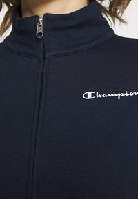 Chaqueta de chándal azul marino con cremallera y alto cuello, con tejido texturizado y un pequeño logo blanco de "Champion" en el área del pecho izquierdo.
